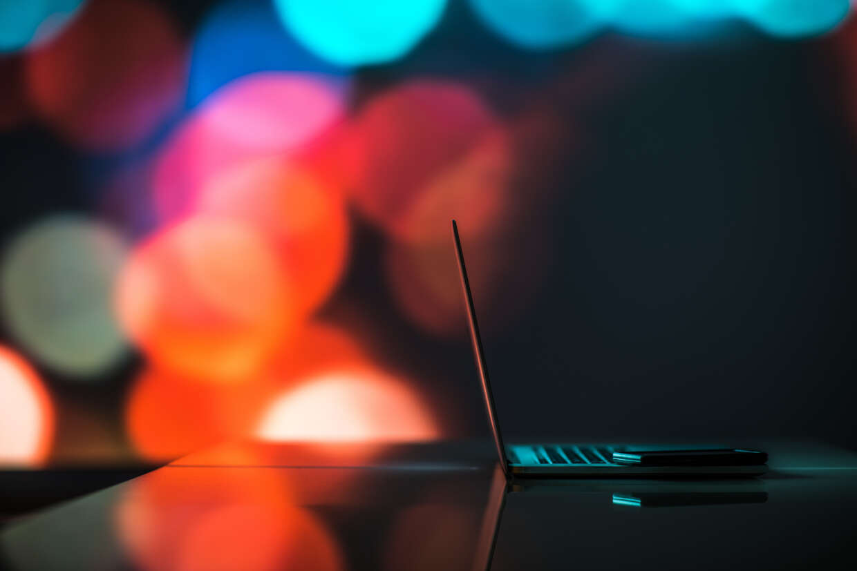 A side profile view of a laptop, resting on a table against a dark background with out-of-focus, vibrant red and orange lights.