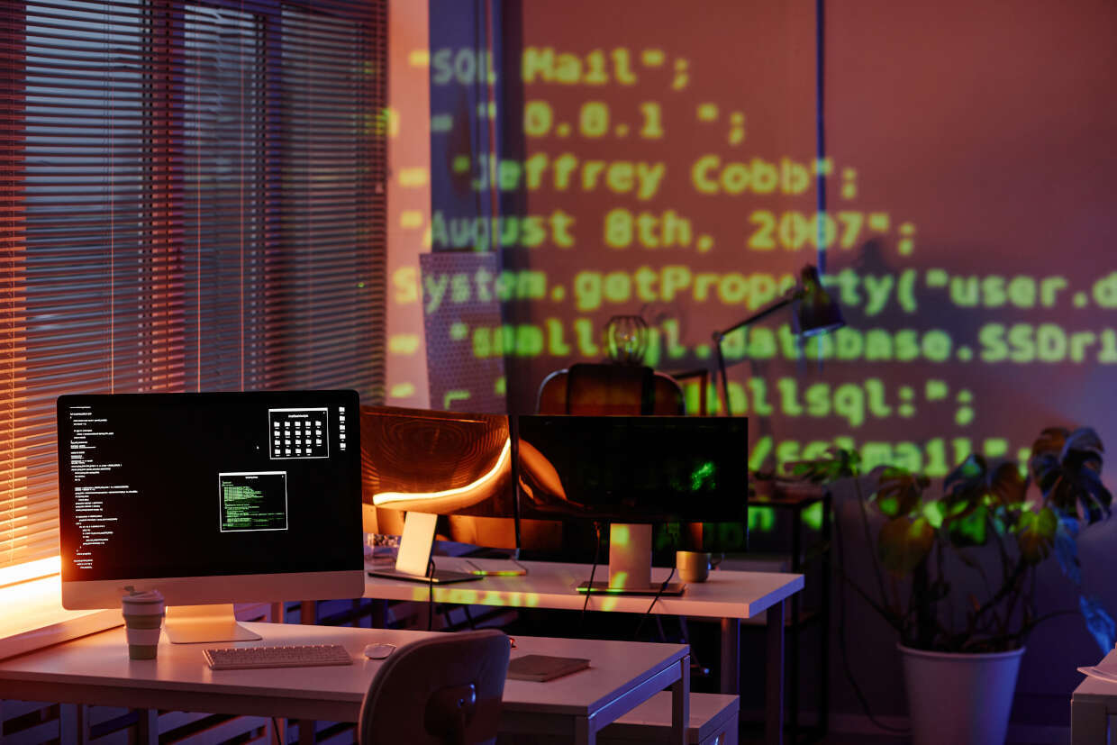 A dimly lit office with two desktop computers on a desk. One screen displays green coding text, while the other shows a system file or code in black and white. The background is illuminated with bright green code projected onto the wall. A plant sits on the desk beside the computers, and a chair is visible in the background. The overall atmosphere feels like a tech or programming environment.
