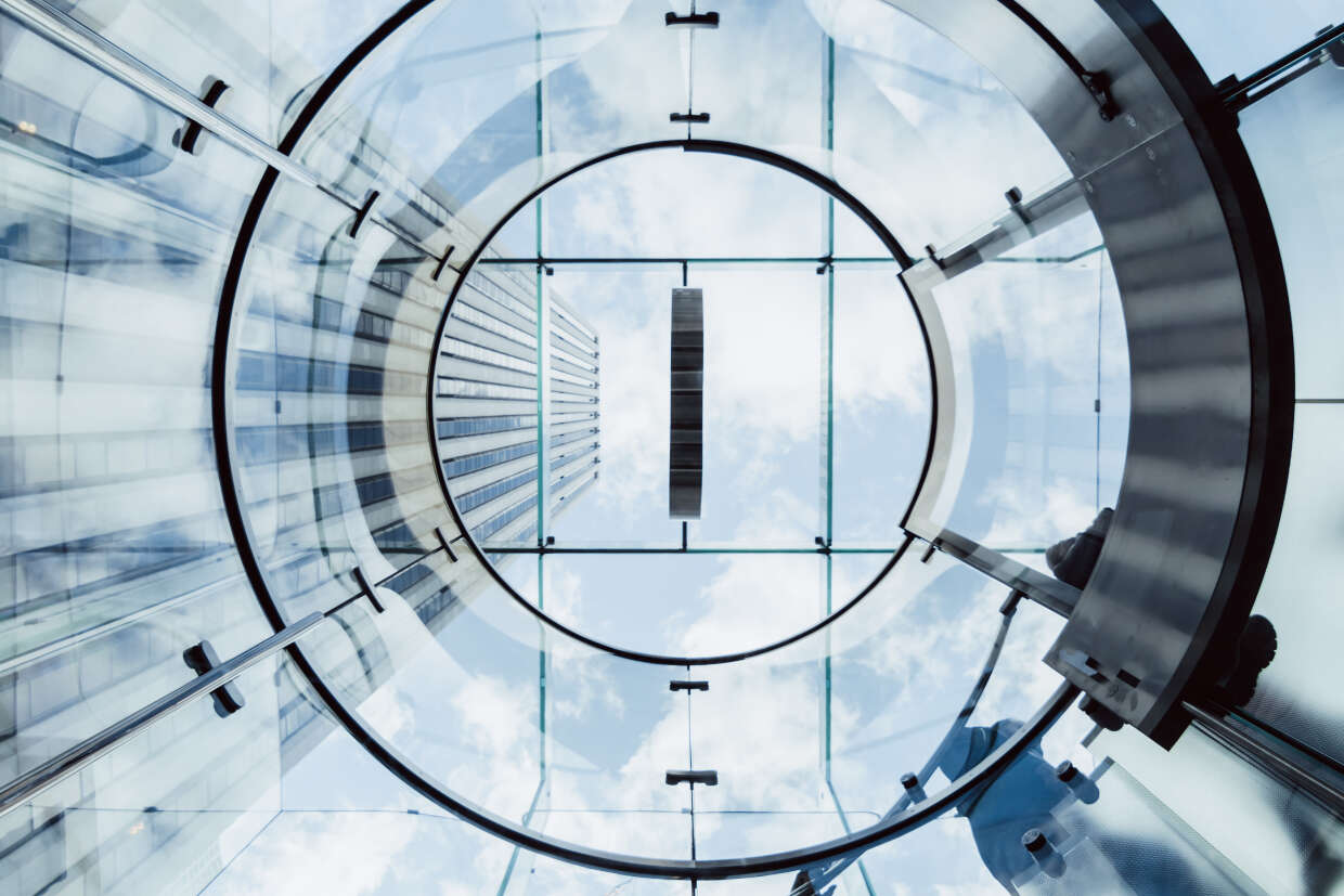 A view looking up through a glass elevator shaft, with reflections of blue sky and clouds. The surrounding buildings are visible through the glass, and metal structural elements frame the circular opening.